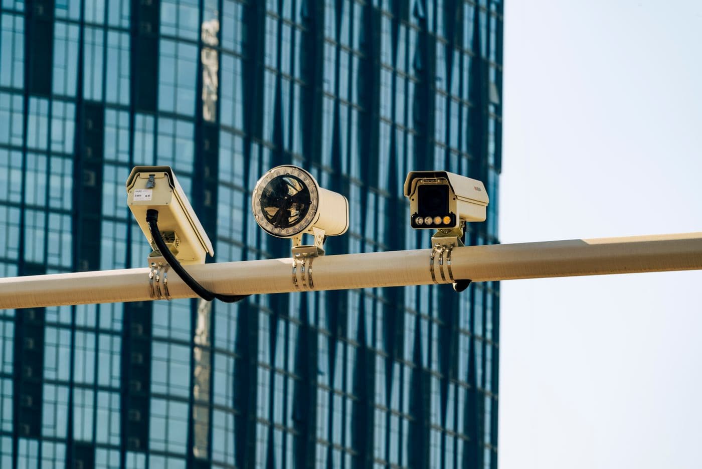 Three surveillance cameras mounted on a bracket in front of a glass tower